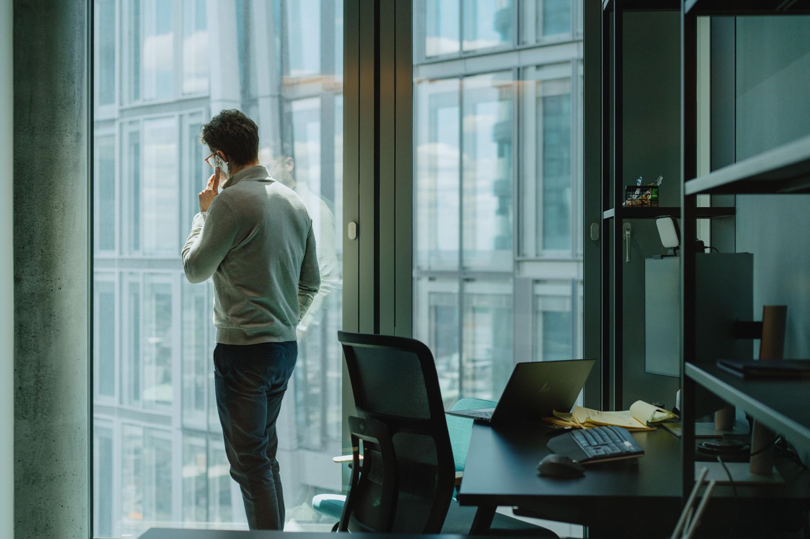 Iemand staat met de rug naar de werkplek bij het raam te bellen, met een leeg bureau en een laptop op de voorgrond